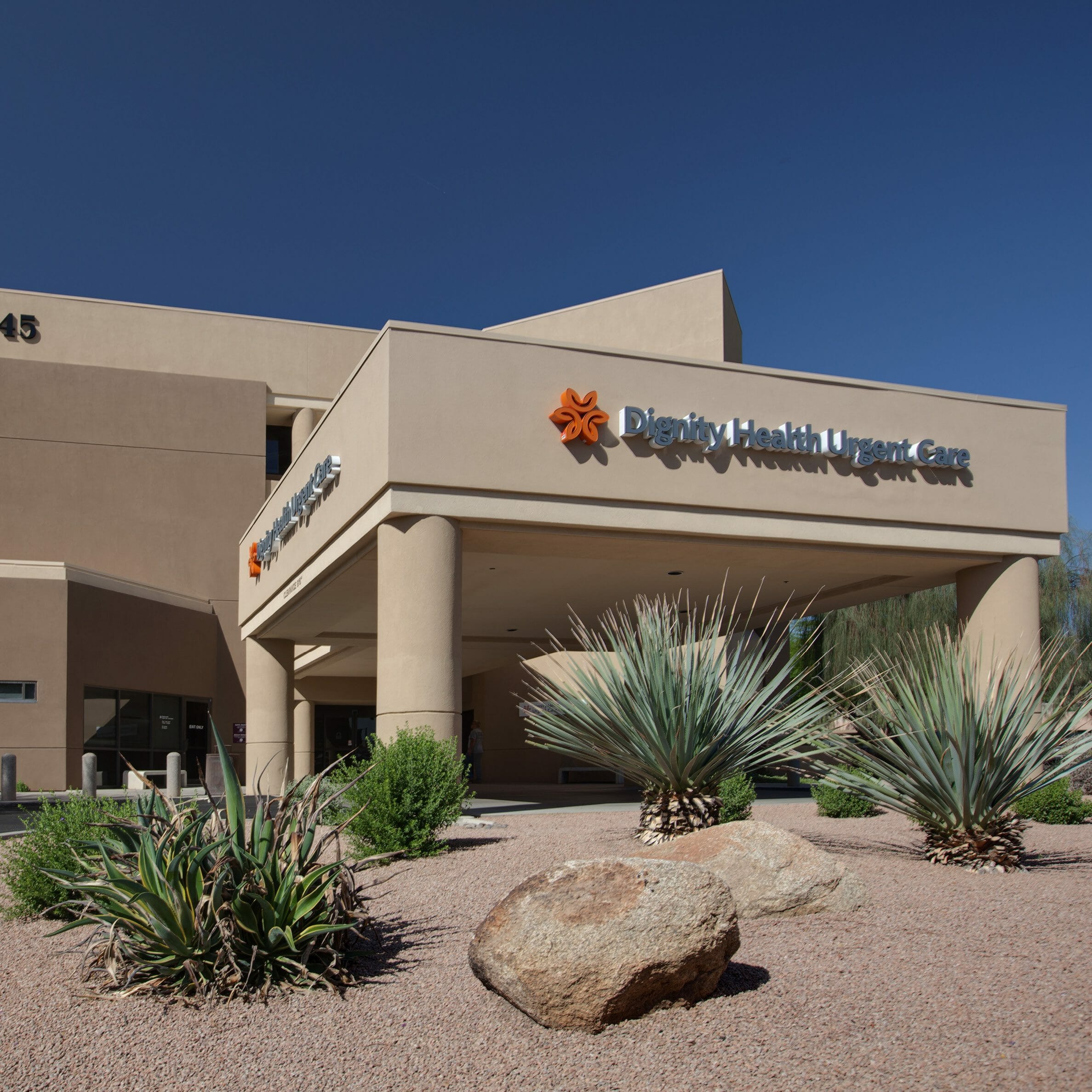 The front of the Dignity Health Urgent Care with a tan, stucco exterior and stucco porte-cochère with desert-like landscape around the building