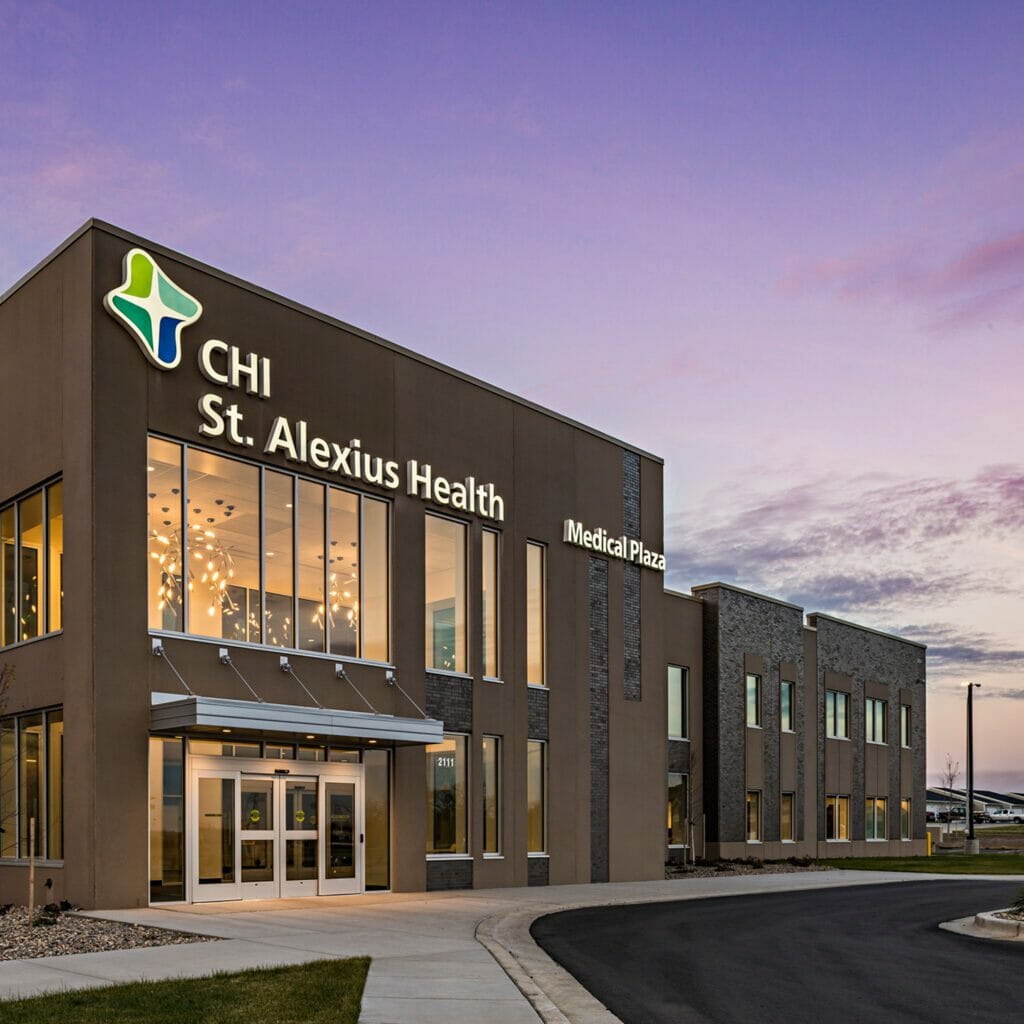 Exterior side angle of front of CHI St. Alexius Health medical plaza at night with stone and stucco finishes and a view of the parking lot