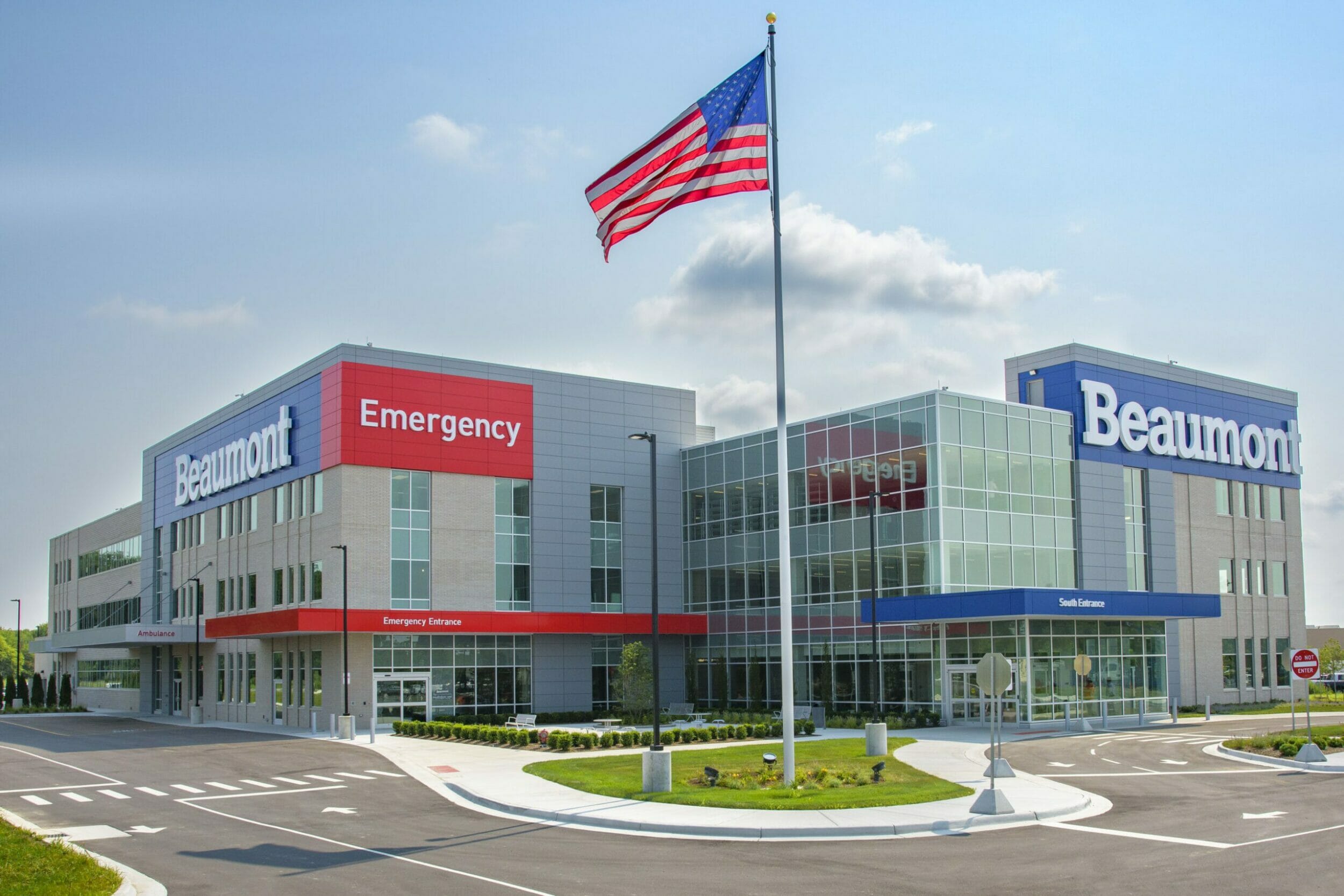 Exterior street view of Beaumont medical center Livonia with a gray exterior, red emergency entrance and blue background feature on building with large, white letters that spells Beaumont