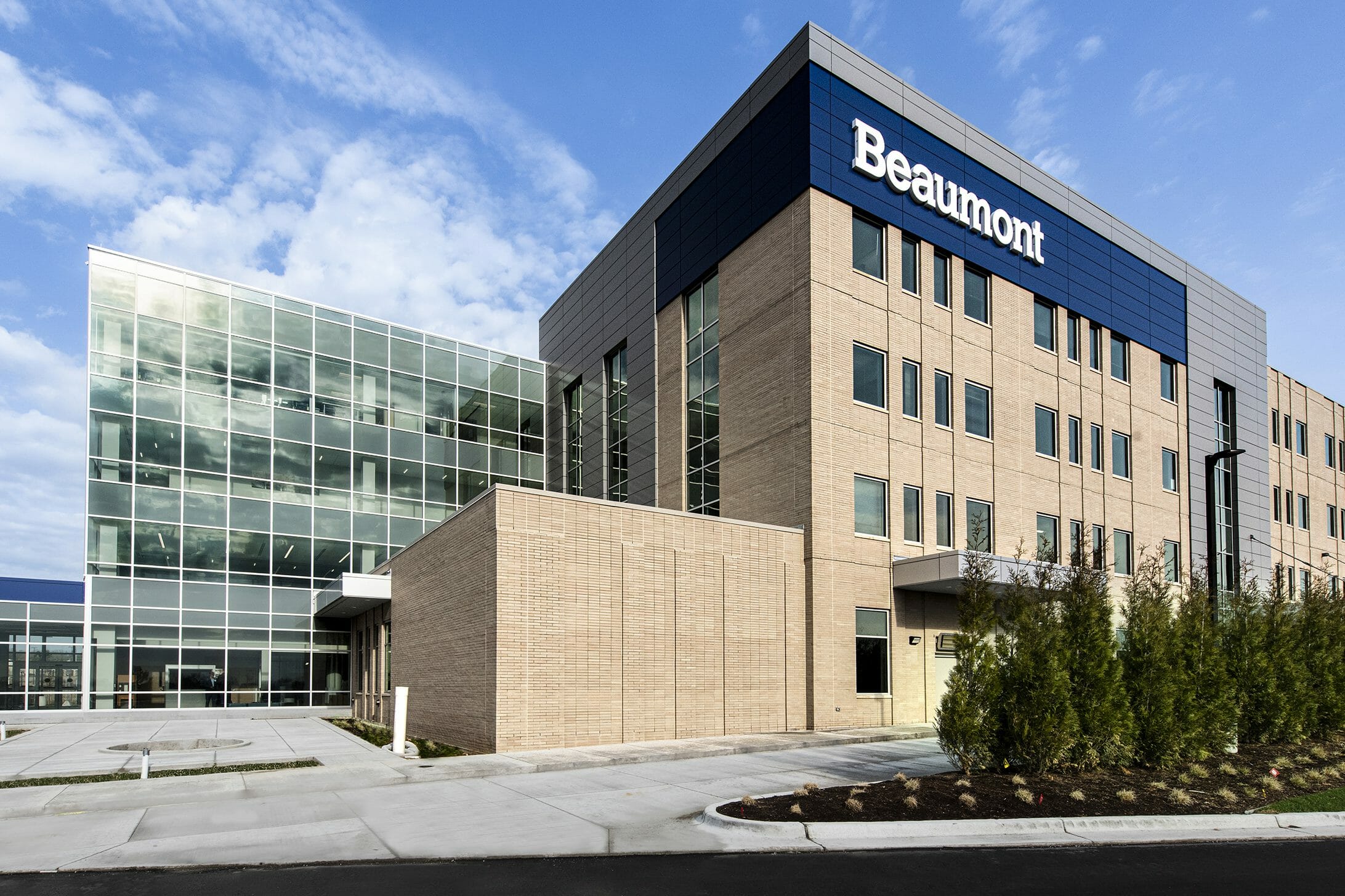 Exterior view of Beaumont medical center with a tan brick, gray, blue, and glass exterior finishes with walking path lined by trees in front of building