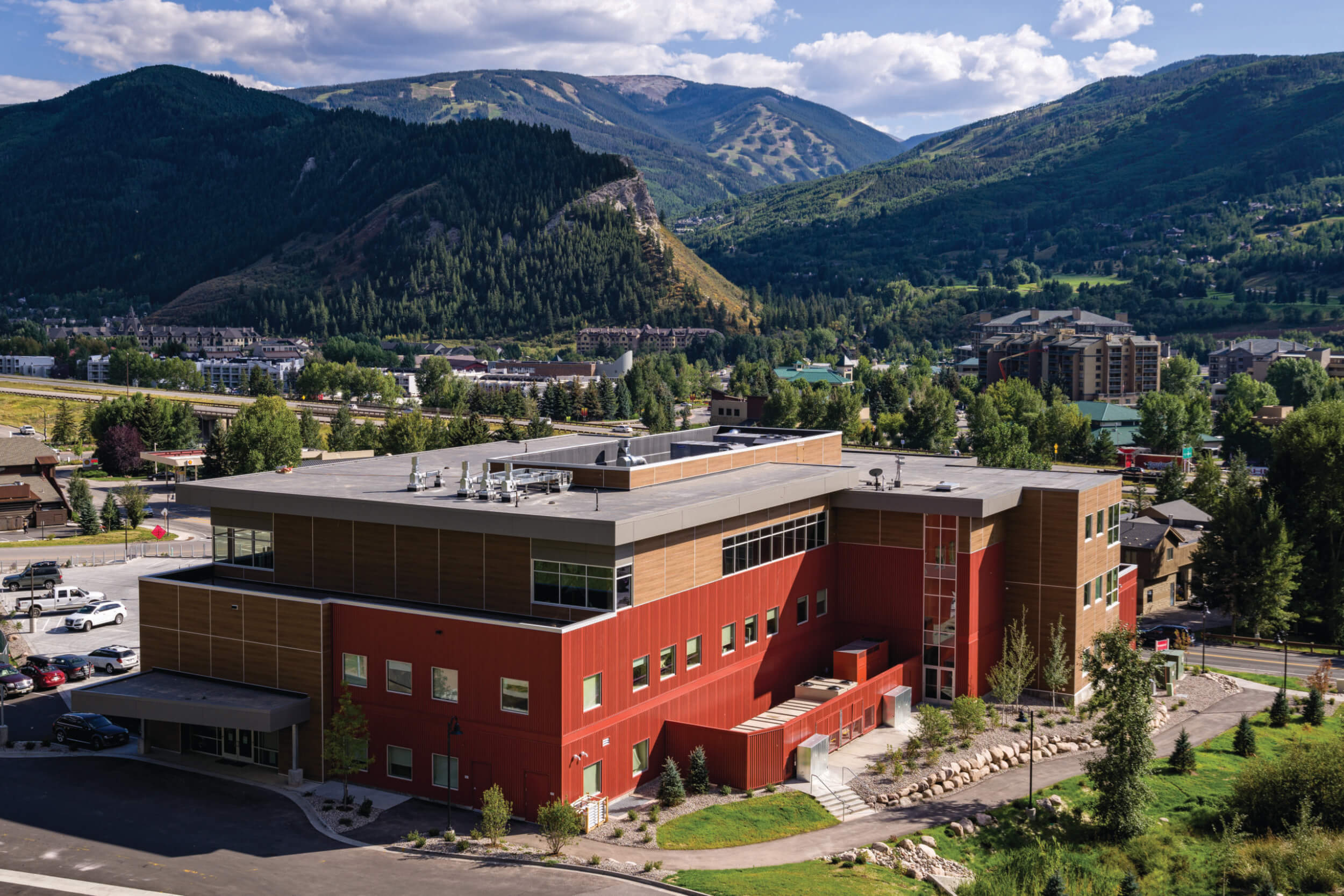 Aerial view of a 3-story medical center with a red and wood exterior and mountains and other buildings in the background