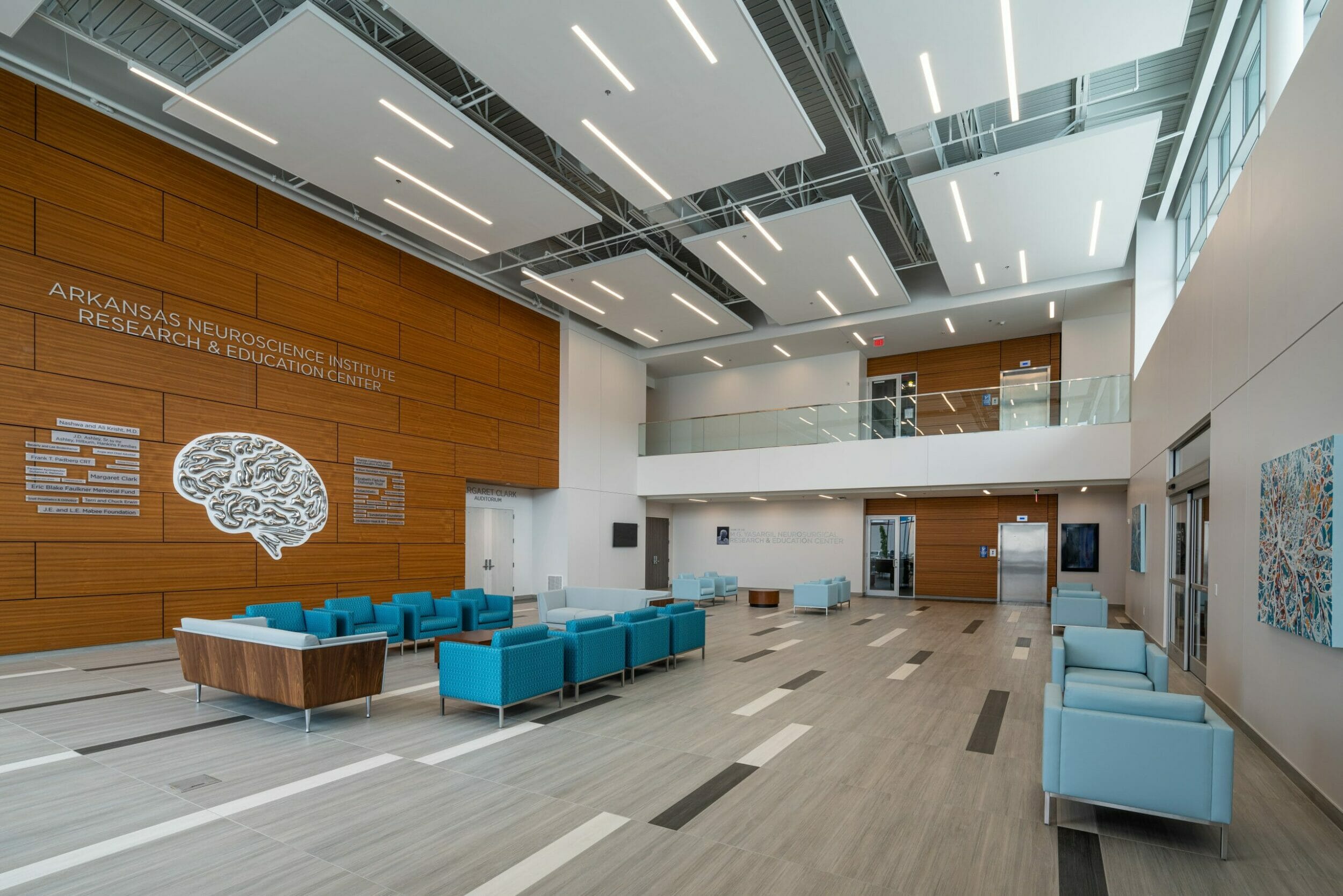 Two-story lobby of the Arkansas Neuroscience Institute Research & Education Center with various wood accents, a large silver brain on a wood wall, and blue furniture