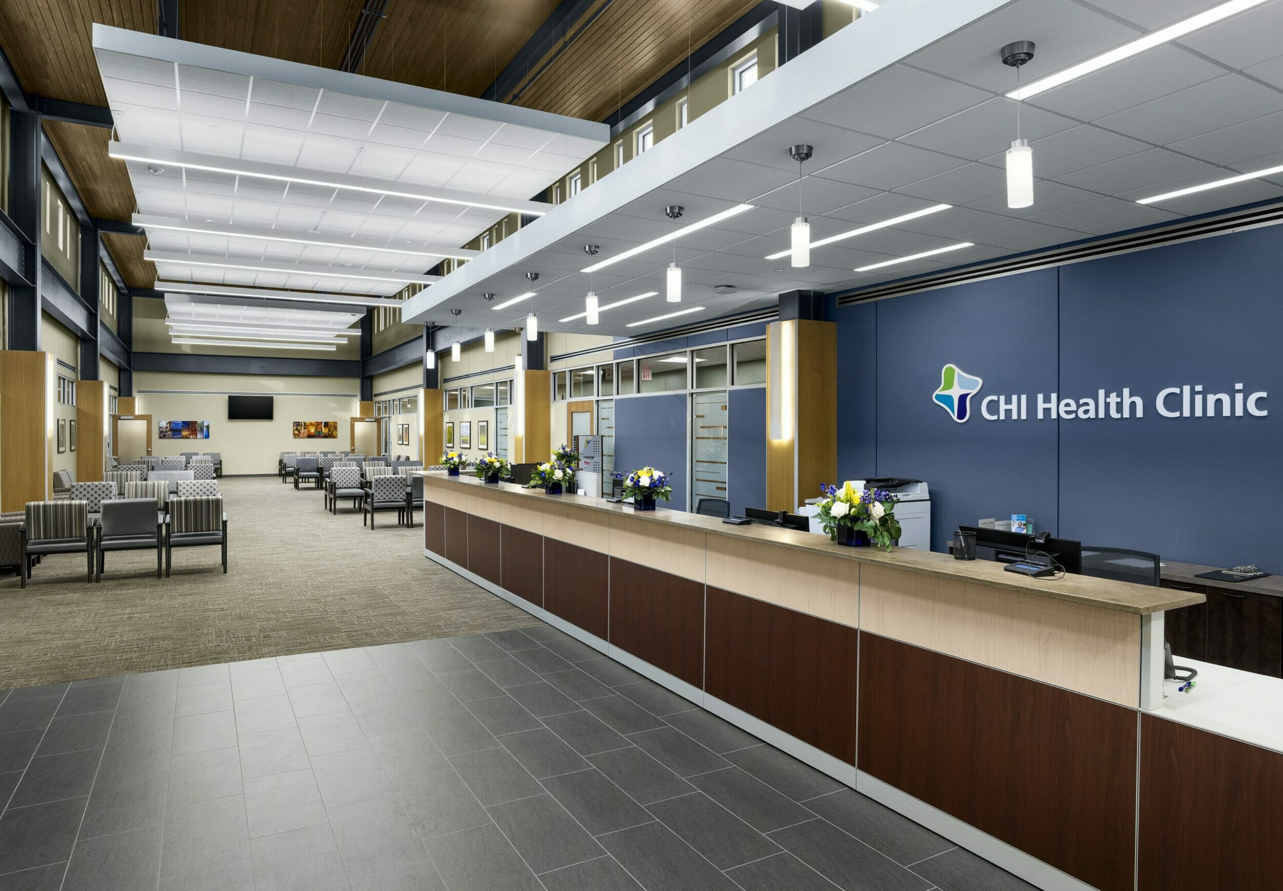 CHI Health Clinic lobby with tiled and carpeted floors, multiple rows of chairs, and a long wood front desk with multiple desks