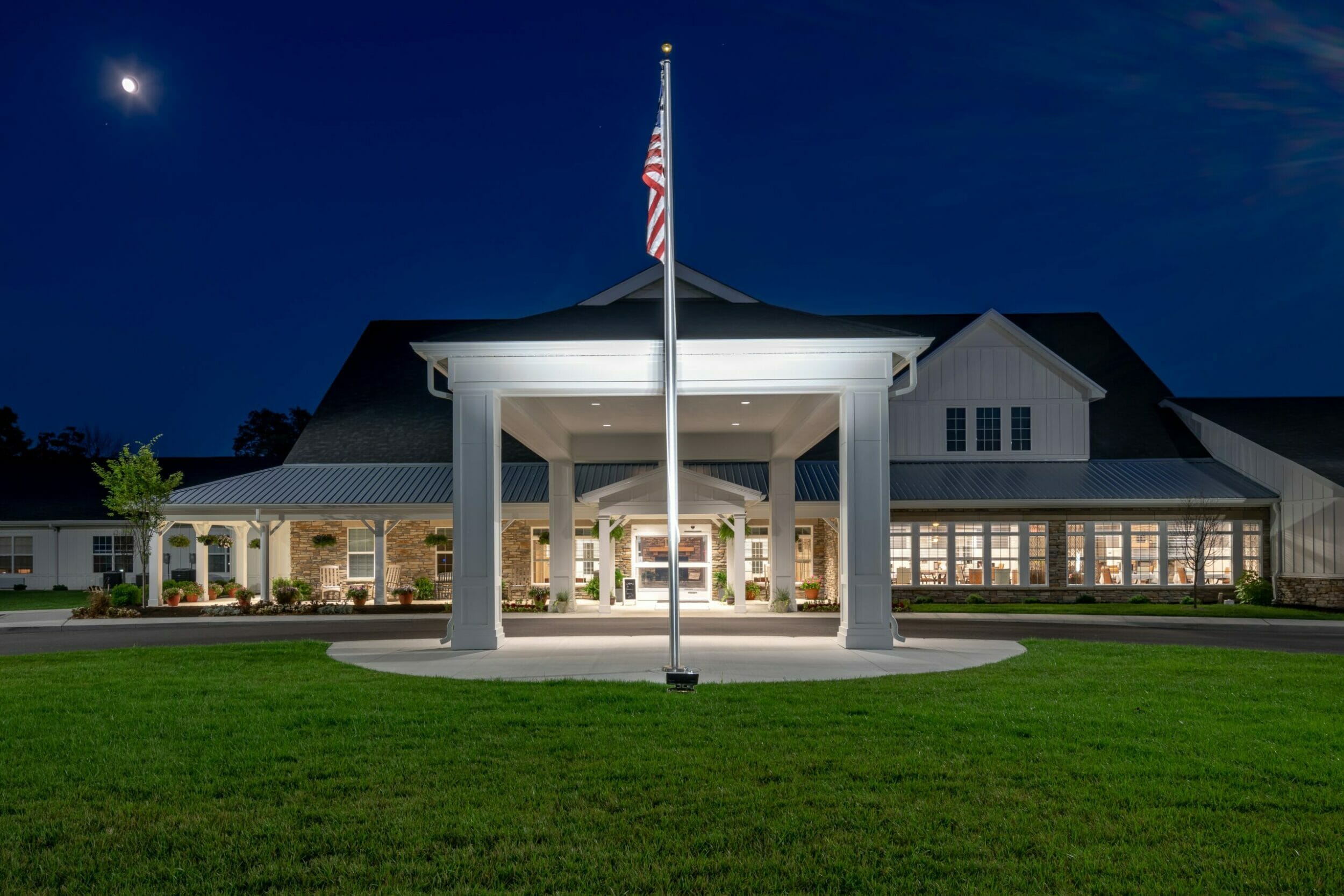 Exterior view of Demaree Crossing senior living at night with the american flag in front of the porte-cochère and front porch with stone accents