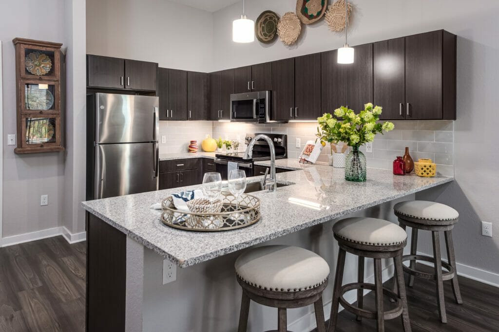Full kitchen with dark brown cabinets, stainless steel appliances, white and gray countertops, and stools for sitting with multiple accessories on the counter
