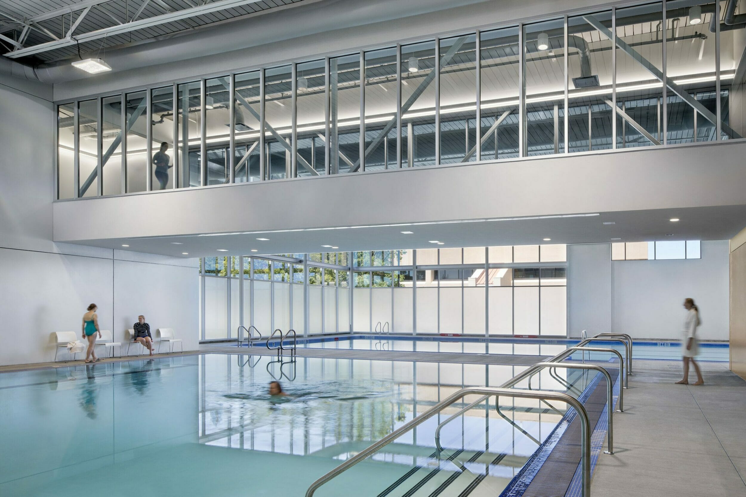 two indoor pools on the first floor of a two-story room inside a wellness center with people swimming and walking above in a glass walkway
