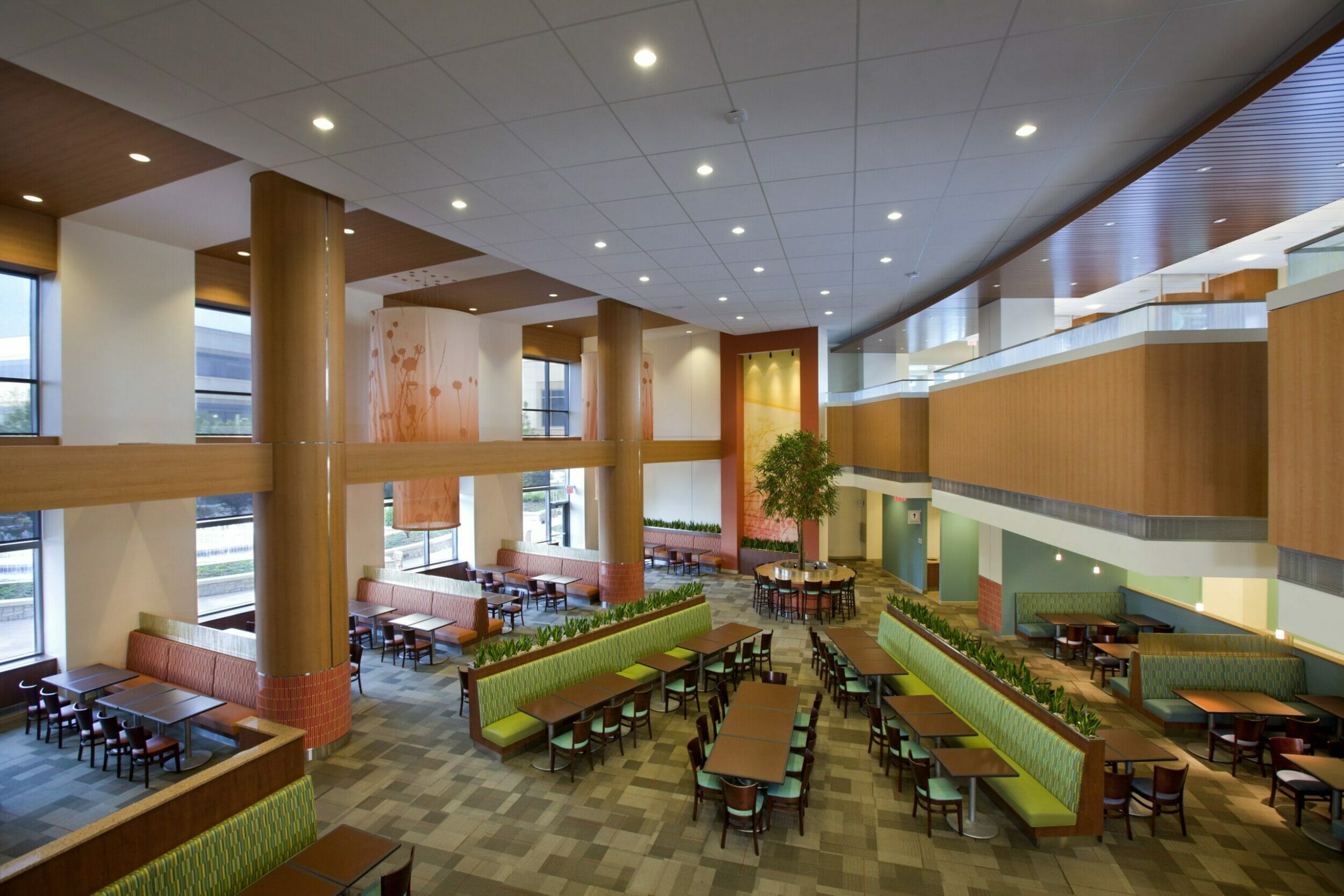 view of cafeteria from upper level. Plentiful seating, large columns, artistic fabric chandeliers, tree and plants