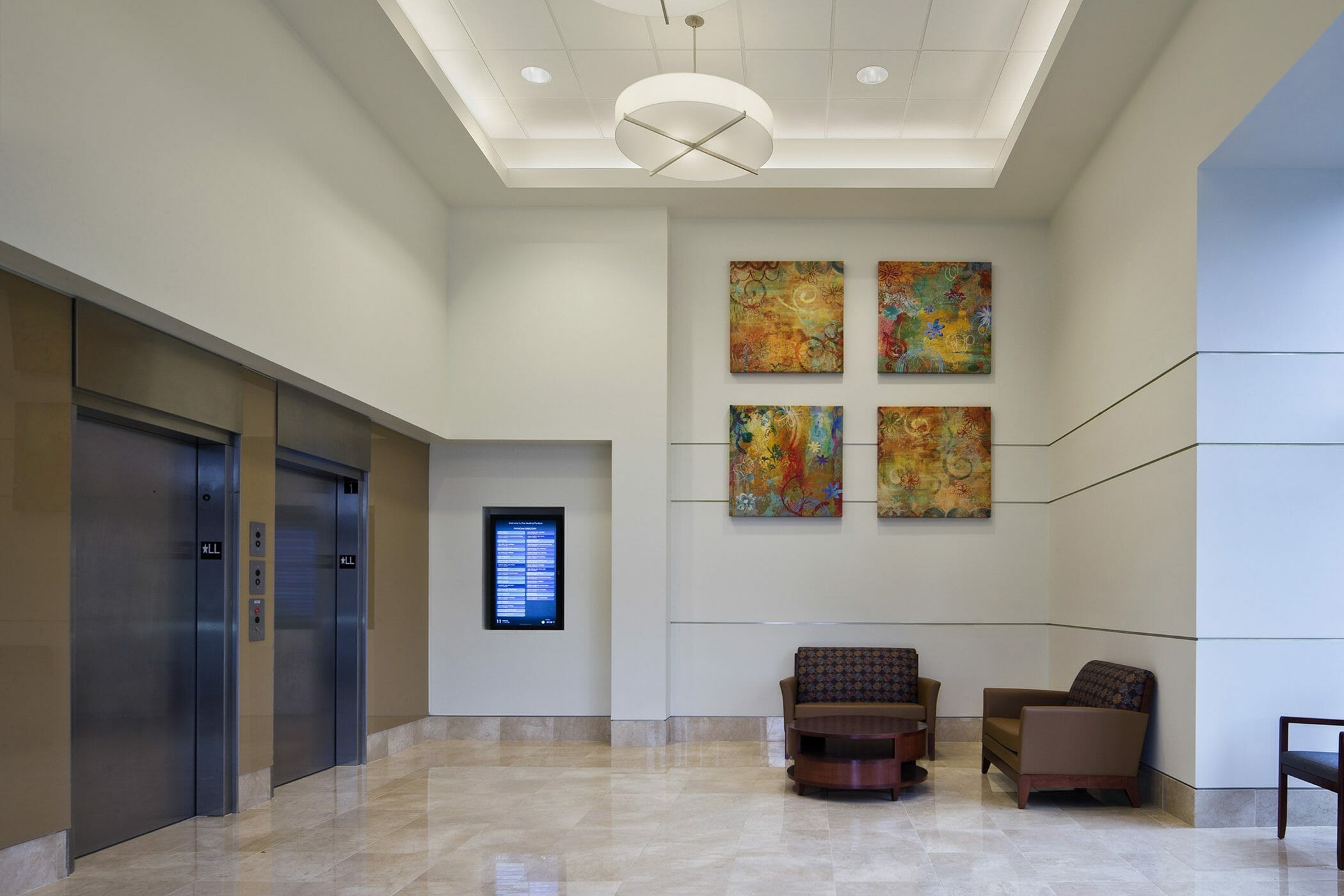 Elevator area on main floor of Angelos Medical Pavilion with tall ceilings and, red chairs, abstract art, and multiple forms of ceiling lights