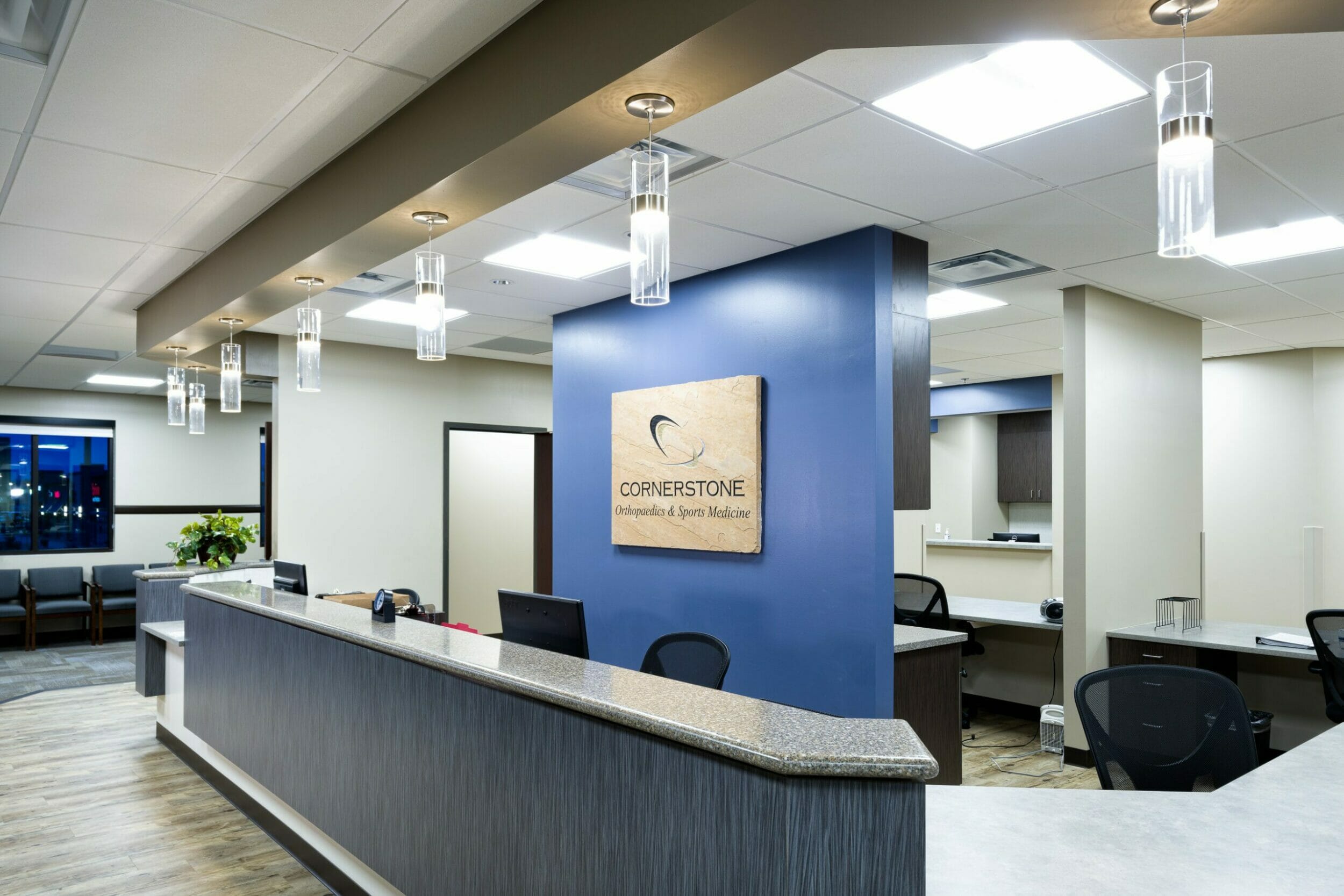 Front desk and lobby inside medical office building with wood and carpeted floors, a long front desk, and blue accents with the Cornerstone orthopedics and sports medicine sign behind the desk.