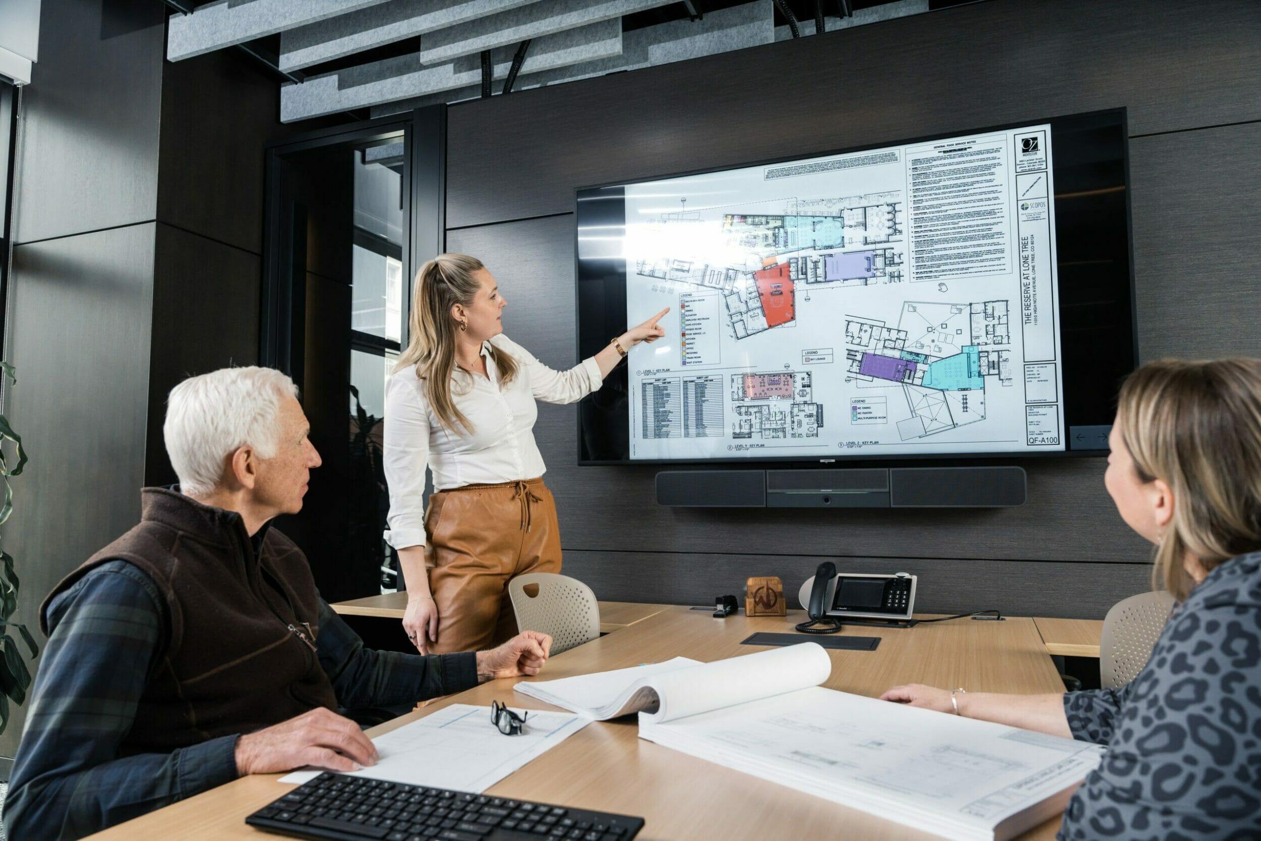 One woman standing next to a screen pointing at blueprints and two other individuals at the conference table looking at the screen