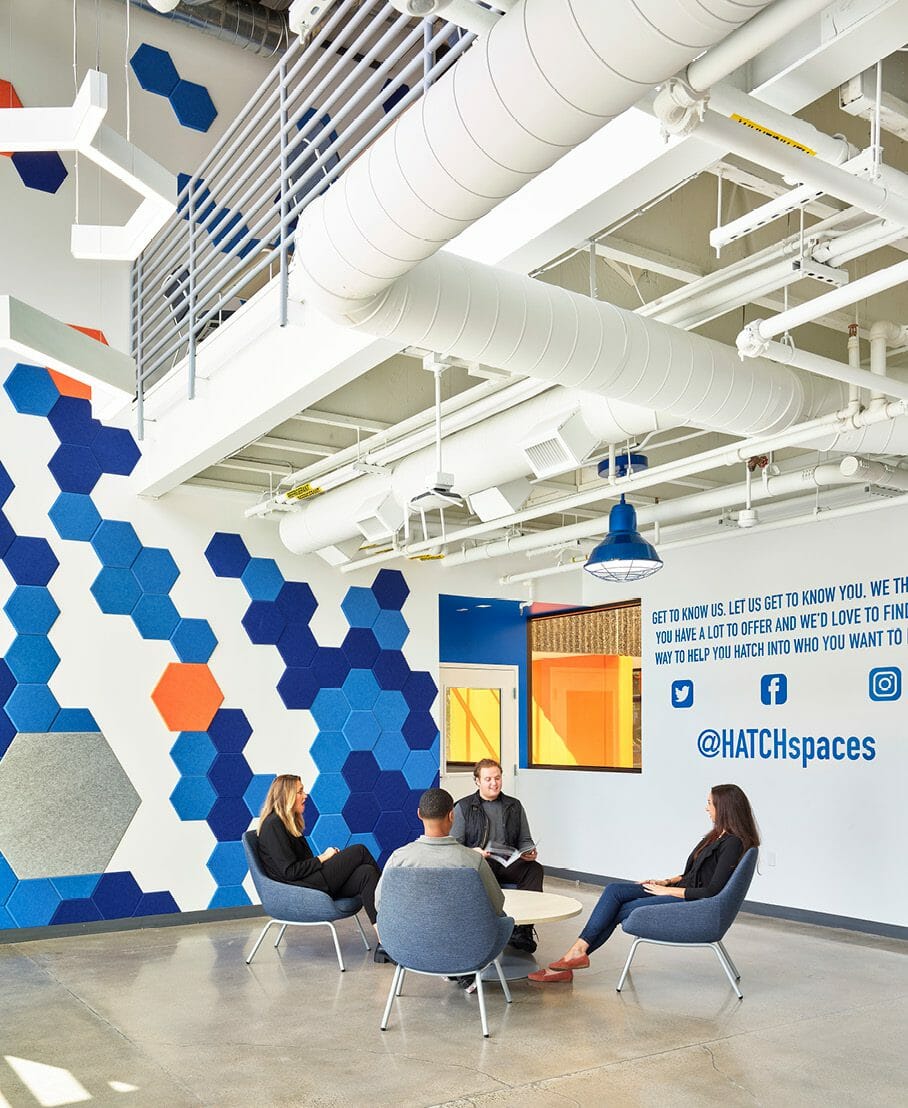 Lobby of HATCHspaces at Irvine Spectrum with people sitting in a lounge area in front of an art wall with blue and orange tiles and a quote wall with social media icons